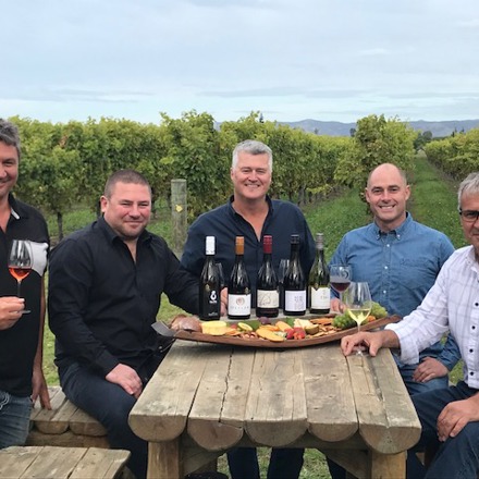 Five men sitting around a table with five bottles of wine and a platter of food being presented
